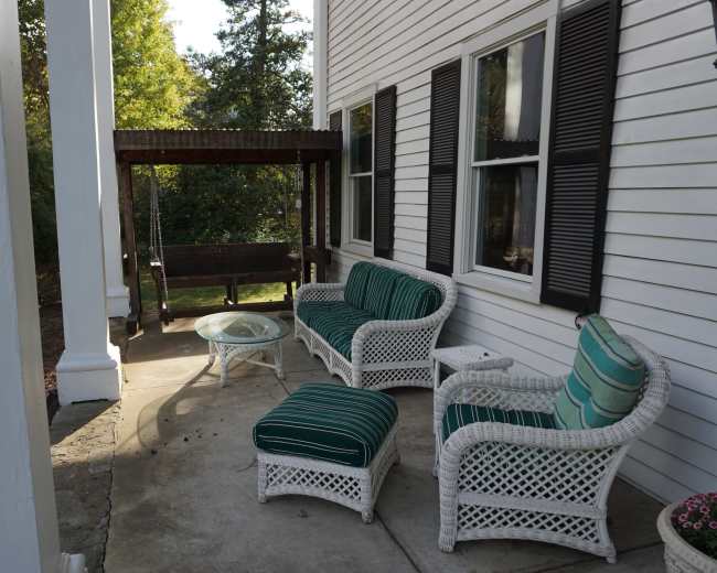 The image shows a porch with white wicker furniture, including a sofa, chairs, and a coffee table, alongside a wooden swing and surrounded by greenery.