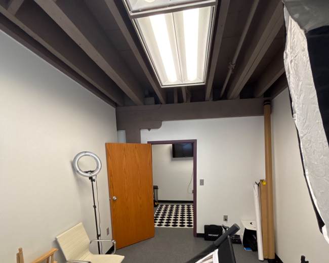 The image shows a small studio space with a chair, a light setup, and a door leading to another room with a checkered floor.
