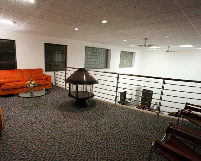 A spacious, well-lit mezzanine features an orange couch, a glass coffee table, and a lamp, overlooking a room with chairs and people.