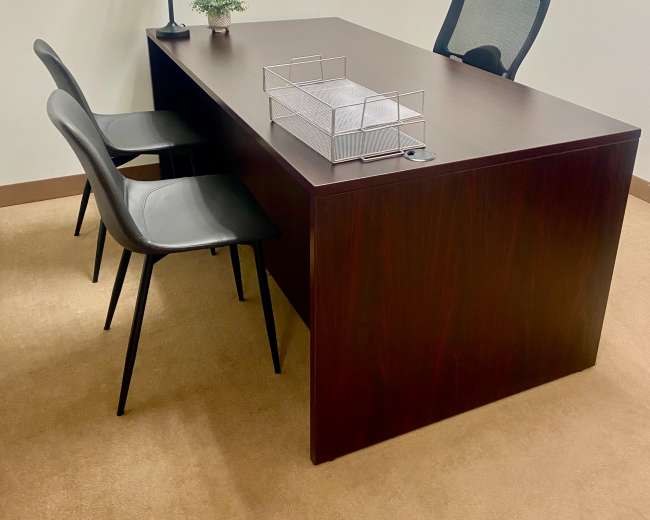 The image shows a minimalist office setting featuring a wooden desk, a lamp, a small plant, three black chairs, and a wire organizer.