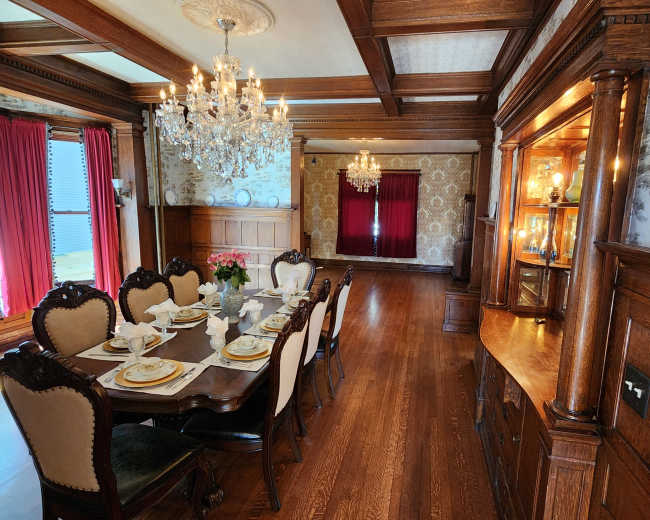 The image shows a formal dining room with a large wooden table set for a meal, surrounded by high-backed chairs, and adorned with chandeliers and decorative wall features.