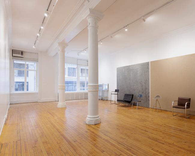 The image shows a spacious, empty art gallery with wooden floors, two large pillars, and minimalist furniture against blank walls.