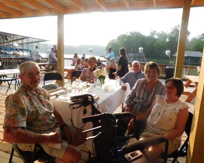 A group of four adults sits at a wooden table on a deck overlooking a body of water, with a festive gathering underway in the background.