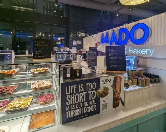 The interior of a bakery named Mado displays various desserts and pastries in a well-lit glass counter.