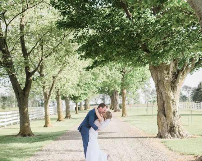 A couple embraces on a gravel path lined with trees and a white fence.