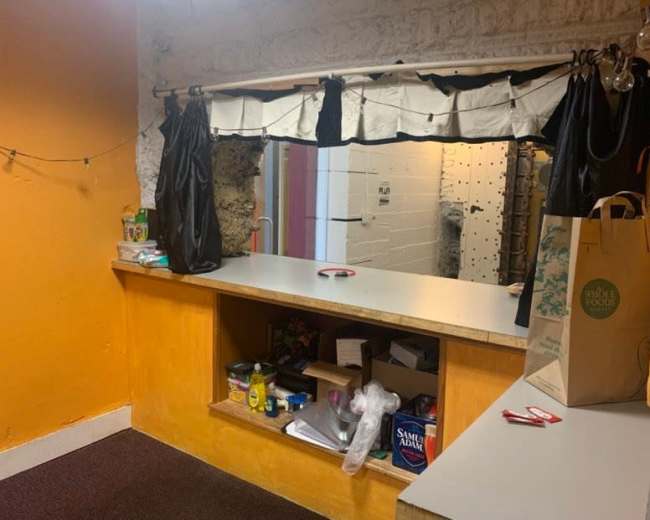 A small room with an orange wall, a counter with various items, and a window covered by black curtains.