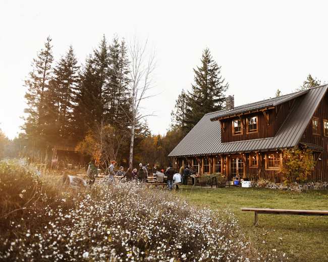 A rustic wooden cabin sits amidst a group of people gathered outside in a natural setting surrounded by trees and wildflowers.