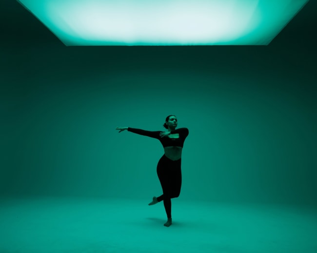 A dancer performs gracefully in a dimly lit studio with an illuminated ceiling casting a green hue.