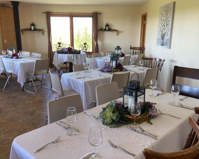 The image shows a dining room with several tables set with white linens, glassware, and floral decorations, ready for a gathering or event.