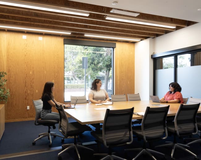 Three women sit at a large conference table in a modern meeting room with a wooden wall and a window.