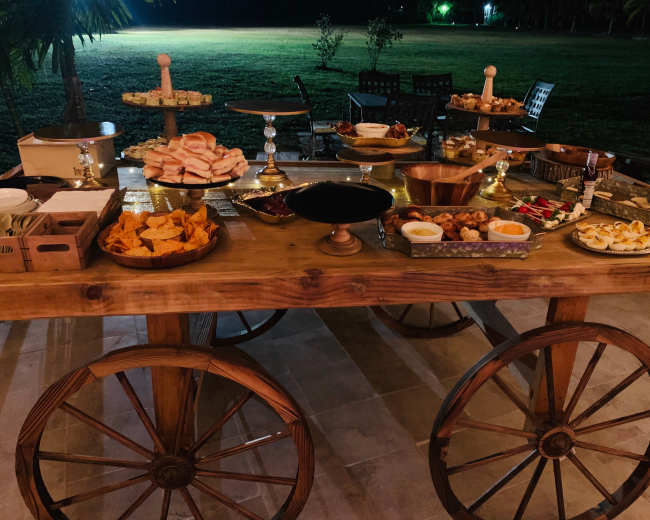 A wooden cart with large wheels displays a variety of food items, including breads, chips, and pastries, set in a dimly lit outdoor area.