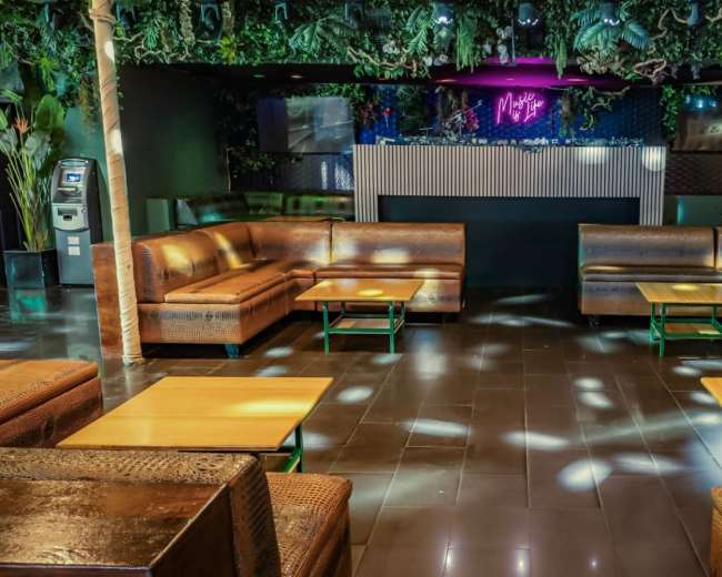 The image shows a modern lounge area with brown leather seating, wooden tables, and greenery overhead, set under dim lighting.