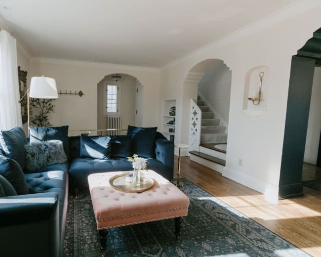 A blue sectional sofa and a pink ottoman are arranged in a bright living room with large windows and a view of a staircase.