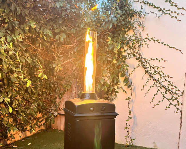 A tall gas flame feature stands on a grassy surface surrounded by greenery.