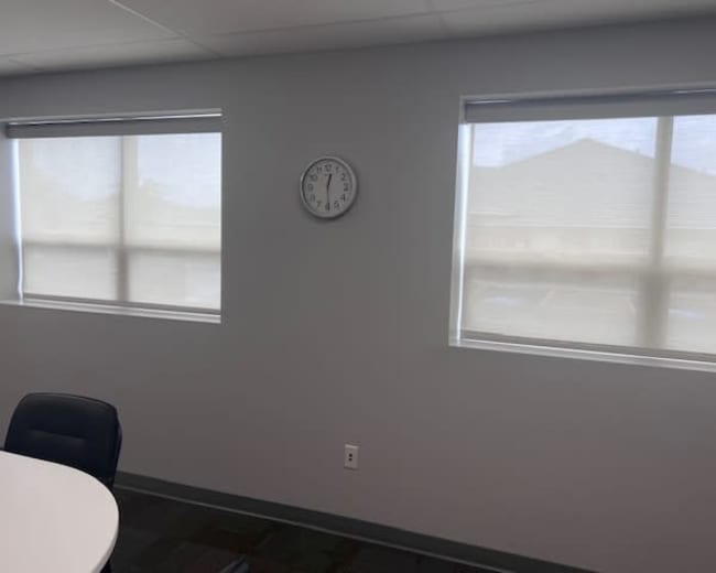 The image shows a minimalist office space with two windows, a clock on the wall, and a round table.
