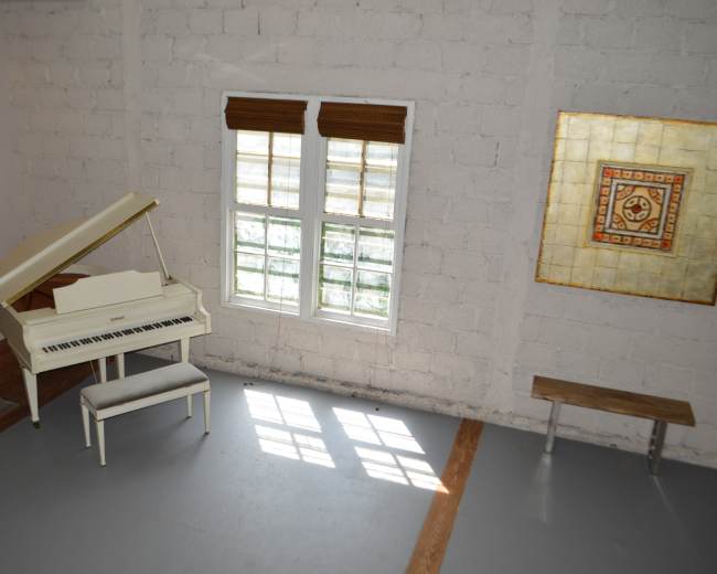 A white grand piano sits beside two large windows in a minimalist room with exposed brick and a bench against the wall.