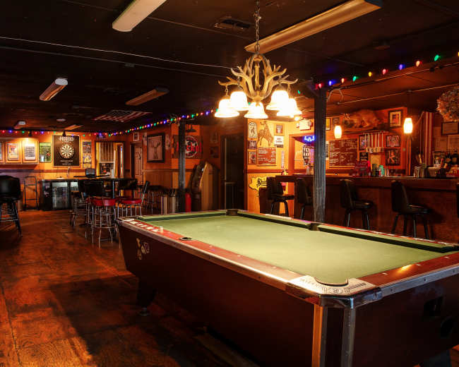 The image shows a dimly lit bar featuring a pool table, colorful string lights, and a wooden counter with stools.