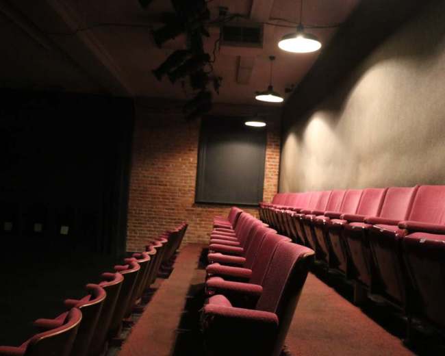 The image shows an empty theater with red seats arranged in rows against a wall, illuminated by overhead lights.