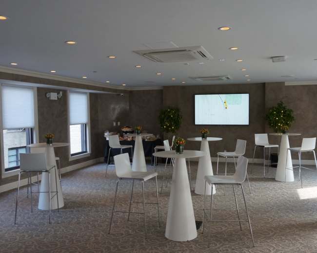 The image shows a modern event space with several high tables and chairs, a large screen on the wall, and decorative plants.