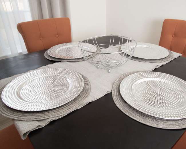 A dining table is set with four silver decorative plates, a woven placemat, and a wire bowl centerpiece, surrounded by two orange upholstered chairs.