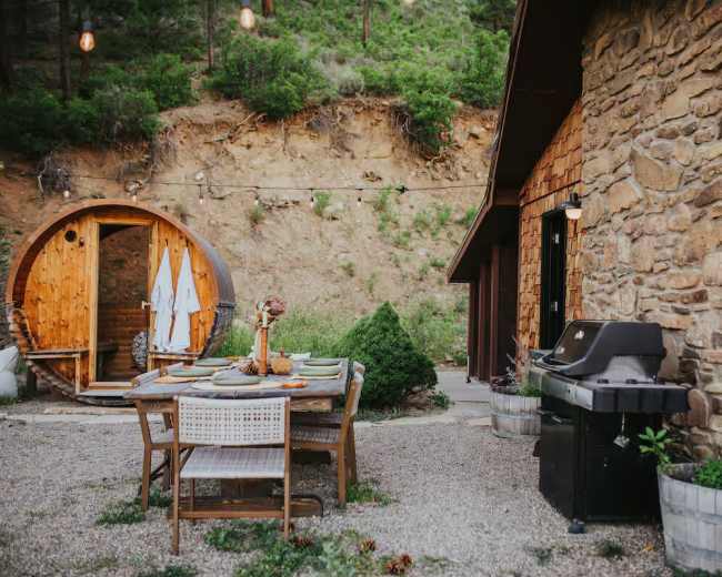The scene features a rustic outdoor dining area with a wooden table set for a meal, a barbecue grill, and a wooden sauna nearby, surrounded by gravel and greenery.