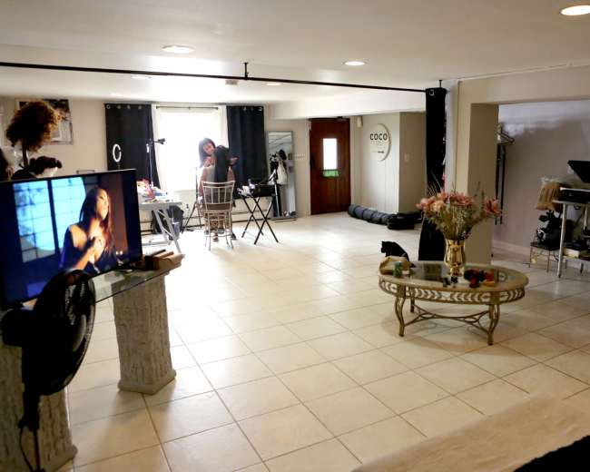 A spacious room with a tiled floor, featuring a lounging area with a television, a dining setup, and a workspace in the background.