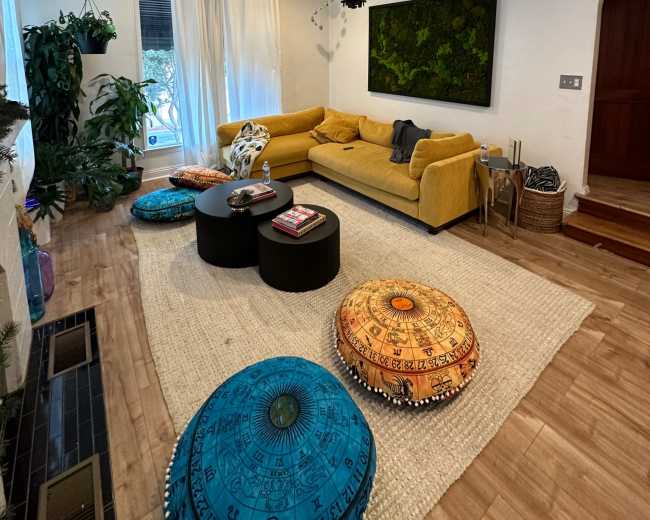 The living room features a yellow sofa, two black circular tables, decorative poufs, and a large wall-mounted art piece, with plants and natural light coming through the windows.