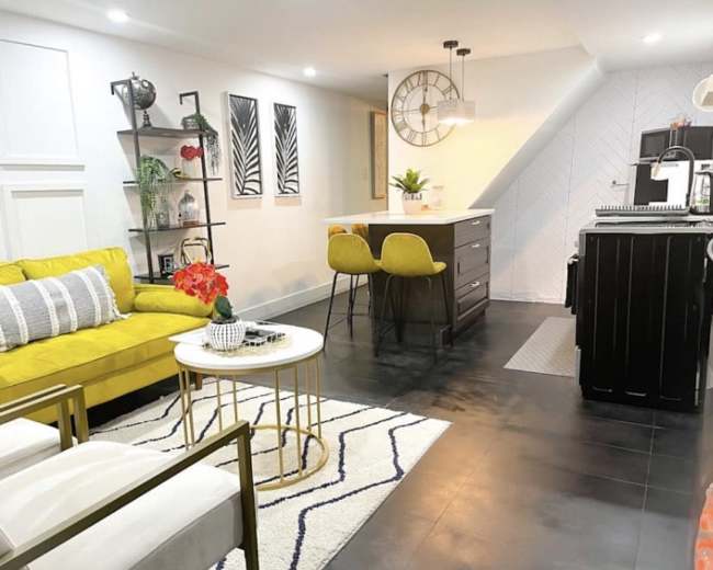 The image shows a modern living area with a yellow sofa, a round coffee table, and a dining area featuring two yellow chairs and a kitchen in the background.