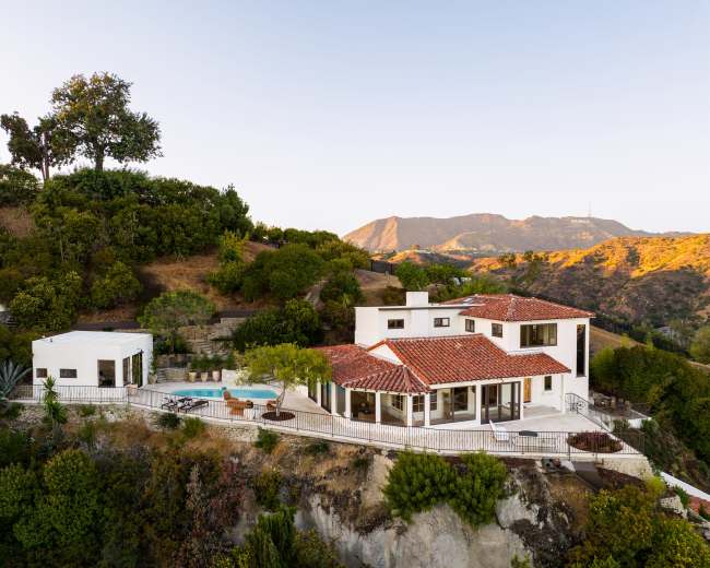 The image shows a modern white house with a red tile roof, surrounded by lush greenery and a pool, set against a mountainous backdrop.