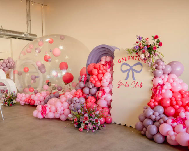 The image shows a festive event space decorated with a large arrangement of pink, purple, and red balloons alongside a sign that reads "Galentine's Girls Club."