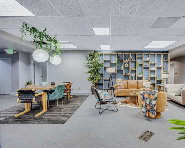 The image shows a modern open workspace featuring a mix of seating areas, a dining table, plants, and decorative bookshelves.