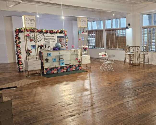 The image shows a spacious, partially decorated room with wooden floors, featuring a decorative display setup on one side and empty furniture pieces scattered throughout.