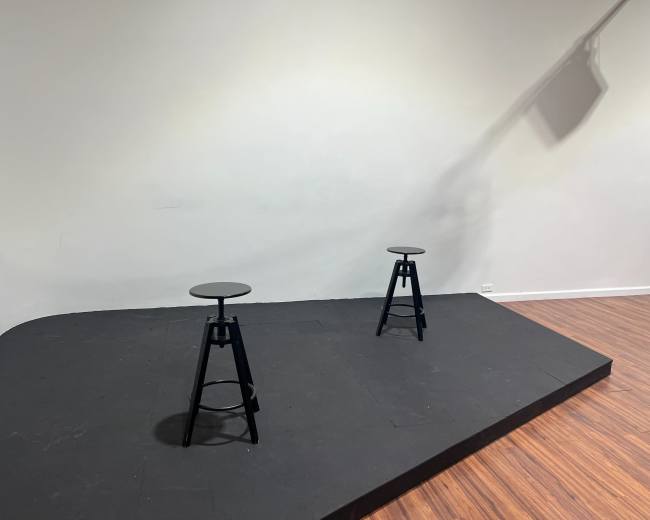 The image shows two black stools positioned on a raised, black stage with a light casting a shadow on the wall.