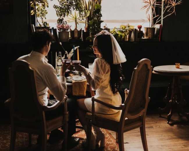 A couple sits at a table adorned with plants, sharing a toast in a sunlit space with large windows.