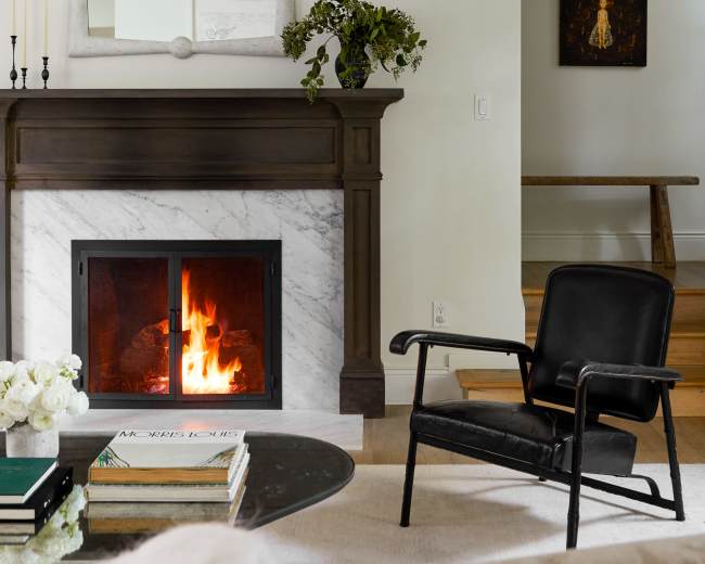 A black chair sits next to a marble fireplace, which has a fire burning and a mirror above it, while a small table with books and a vase of flowers is in the foreground.