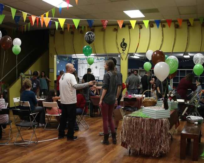 The image shows a lively indoor gathering with colorful balloons, banners, and decorations, where people are interacting at tables filled with party supplies.