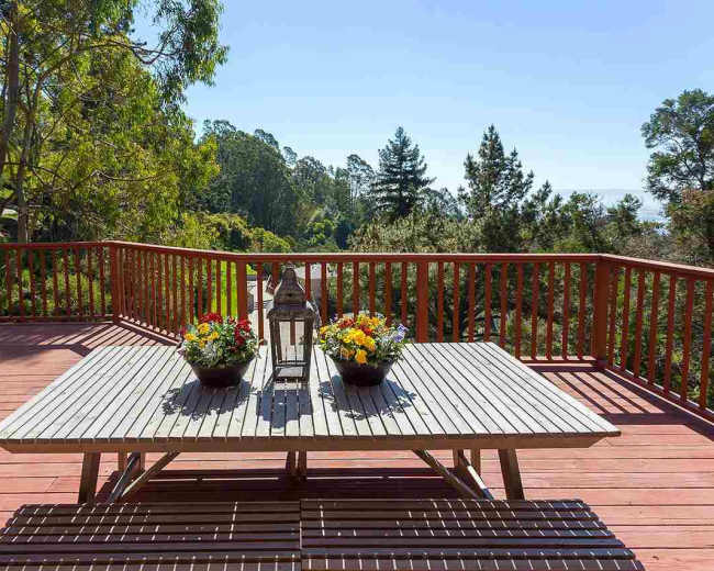 A wooden deck features a table adorned with flower pots and a lantern, overlooking a lush green landscape.