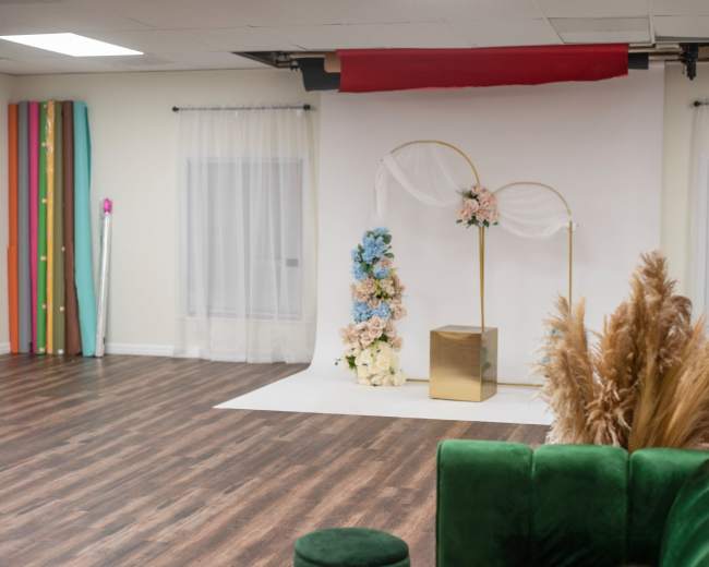 A spacious event room features a white backdrop adorned with floral arrangements, a gold arch, and a square pedestal, with colorful decorations along the sidewalls.