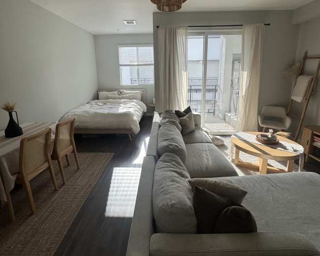 The image shows a modern studio apartment featuring a bed, a seating area with a couch, a dining table, and large windows with curtains.
