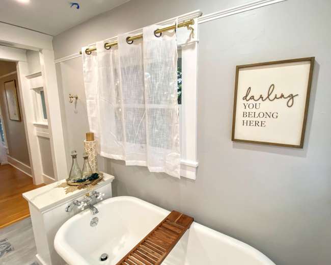 A bathroom with a vintage bathtub, light curtains over a window, and a framed sign on the wall that reads "darling you belong here."