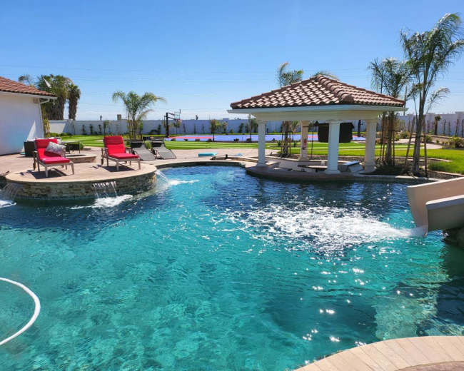 The image shows a landscaped pool area with a gazebo, lounge chairs, and palm trees under a clear blue sky.