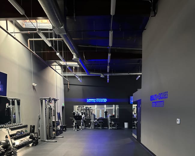 The image shows a spacious gym interior with exercise equipment arranged along the walls and a mirrored area at the far end.