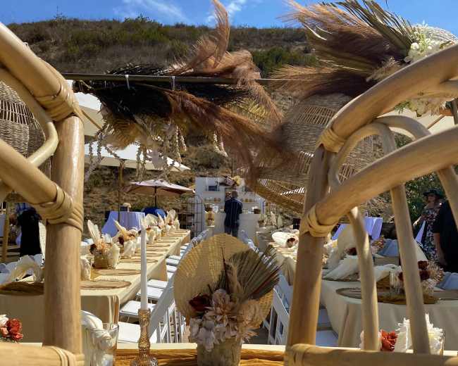 A set of empty bamboo chairs facing a decorated outdoor dining area with tables adorned in neutral tones and floral centerpieces.