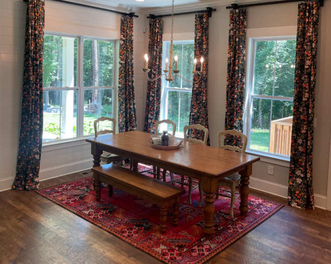 A dining room features a wooden table surrounded by chairs, with floral curtains framing large windows that overlook a green backyard.