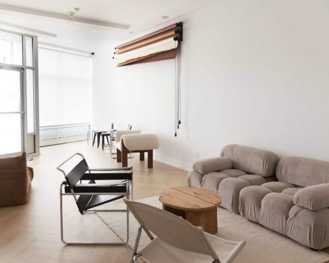 The image shows a modern, minimally furnished interior space with various seating arrangements, including a large couch, armchairs, and small tables.