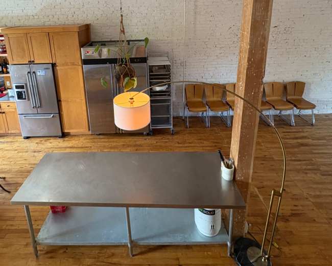A minimalist kitchen features a metal table, wooden cabinetry, and a hanging light above, with a waiting area of chairs in the background.