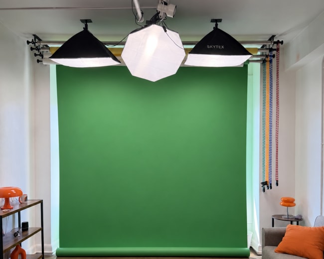 The image shows a studio setup featuring a green backdrop, three lighting fixtures, and a sofa with an orange pillow.