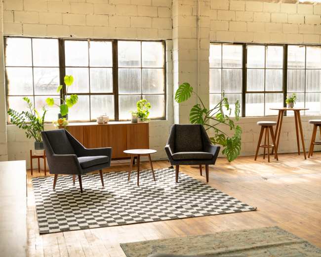 A modern living space features two upholstered chairs, a small table, and a bar-style table arrangement, all surrounded by large windows and plans.