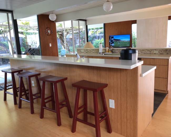 A modern kitchen with a bar counter and four wooden stools, featuring a television and windows that overlook a garden area.