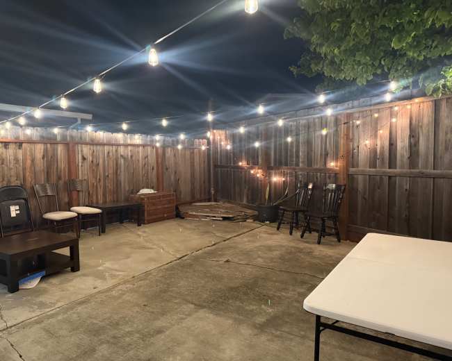 A patio area is illuminated by string lights, featuring various seating options and a few wooden structures against a wooden fence.
